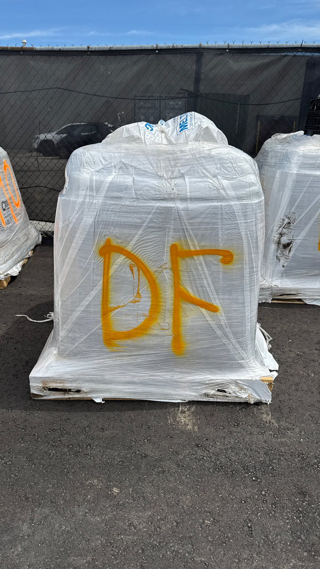 A large white plastic-wrapped palletized load marked with yellow spray-painted letters 'DF', photographed outdoors on a paved surface with other wrapped loads and a fence in the background.