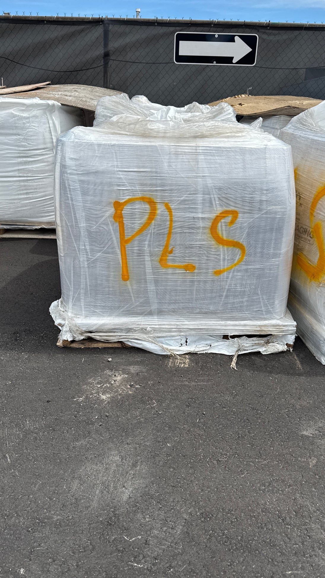 A large white palletized load wrapped in clear plastic, marked with yellow 'PLS' lettering, sitting on asphalt outdoors.