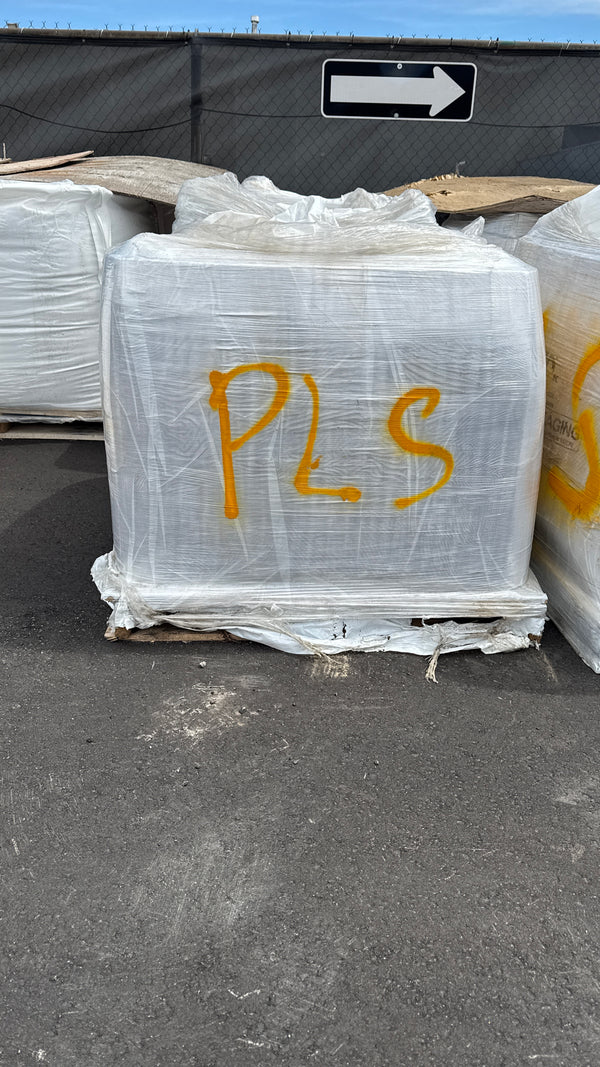 A large white palletized load wrapped in clear plastic, marked with yellow 'PLS' lettering, sitting on asphalt outdoors.