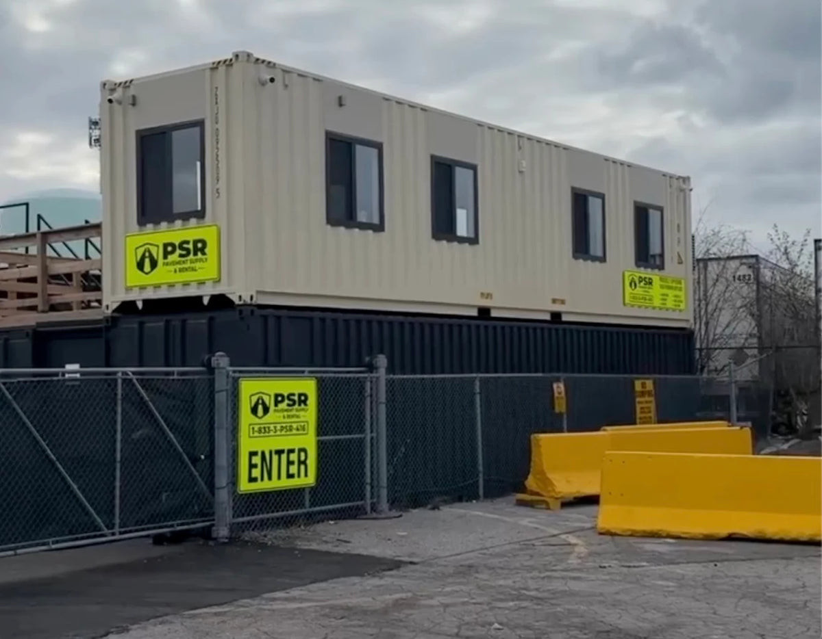 A two-story modular office container home with a cream-colored top floor and a dark gray bottom floor, featuring yellow PSR safety signs, a chain-link fence, and yellow barriers in an outdoor industrial setting.